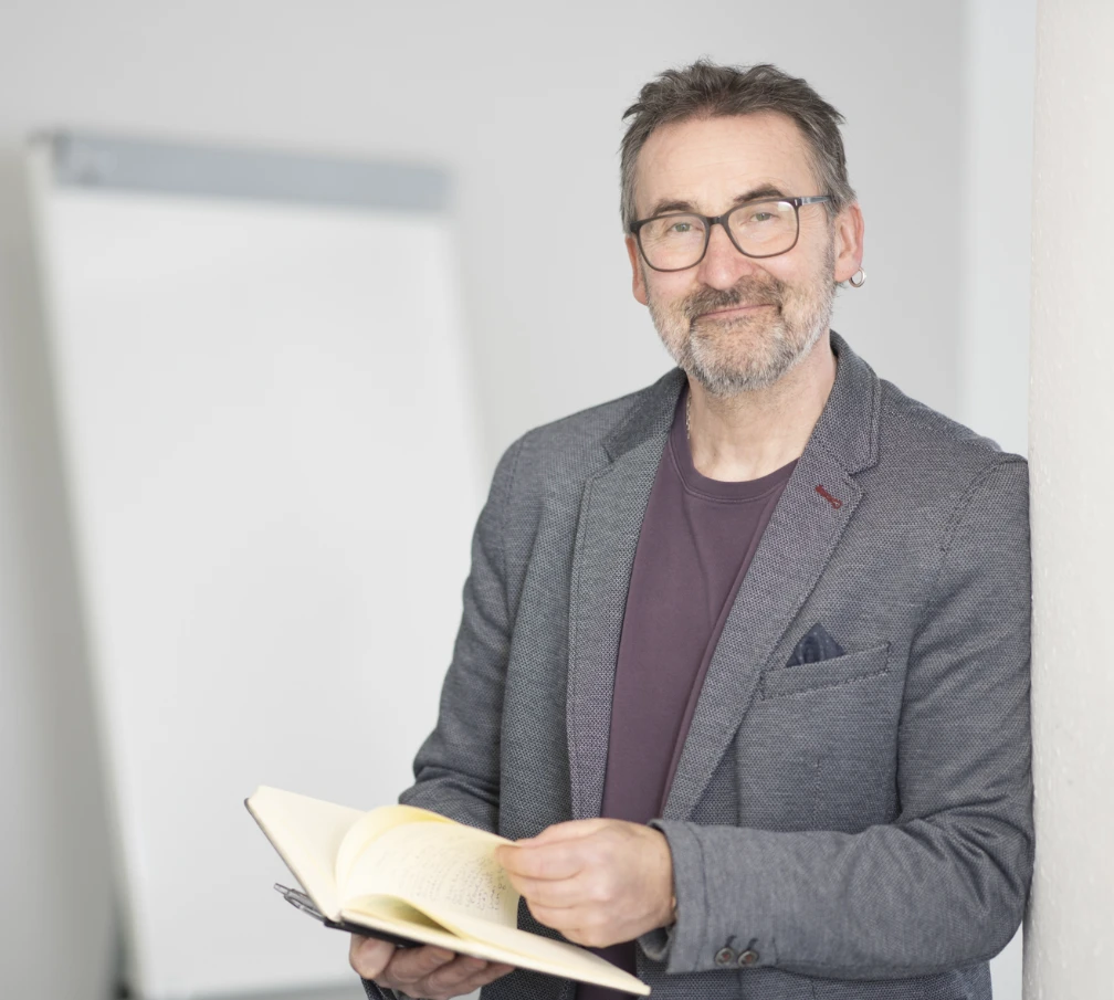 Nachhaltigkeitsberater Timothy C. Vincent mit einem aufgeschlagenen Notizheft in der Hand. Im Hintergrund ist unscharf ein Flipchart zu sehen.